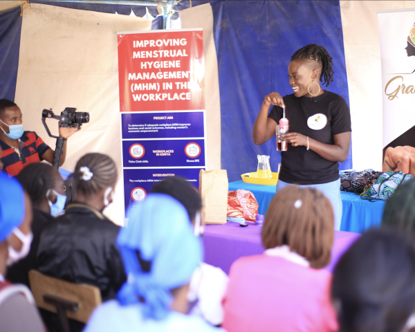 A smiling woman stands in front of an audience giving a demonstration on how to use a menstrual product. 